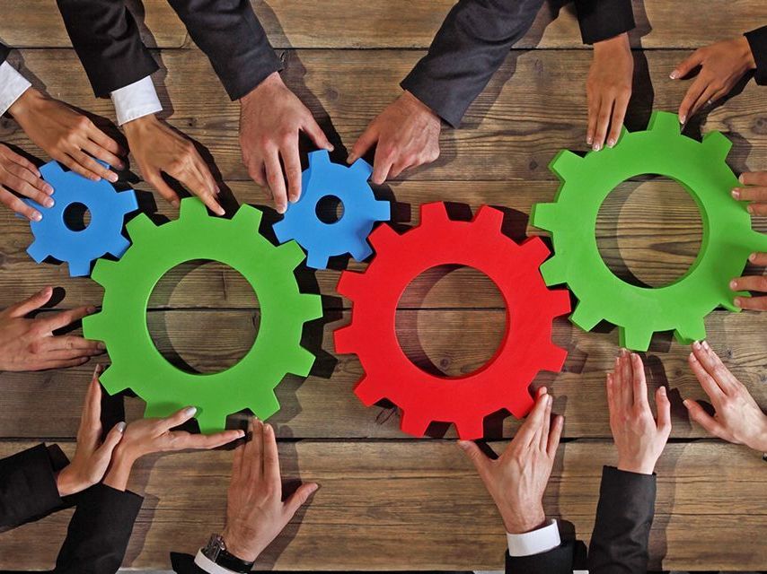 Blick von oben auf einen Tisch, auf dem fünf Zahnräder liegen. Um den Tisch herum sitzen Acht Männer in Anzügen.